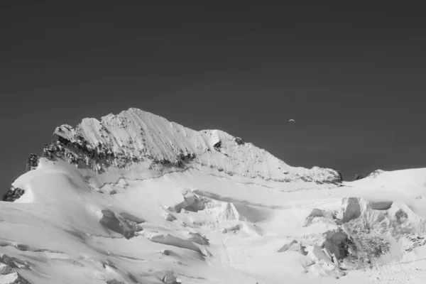 Photographie de montagne Alpes — la Barre des Écrins dans le massif des Écrins, avec un parapente minuscule dans le ciel, en noir et blanc, série Neige & Roche par Adelin Benard..
